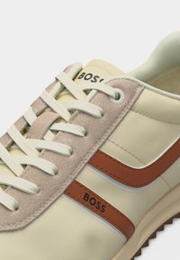 Κοντινή λήψη αθλητικού παπουτσιού σε κρέμ και μπεζ χρώμα με λευκά κορδόνια και καφέ ρίγα που φέρει το όνομα της μάρκας "BOSS" στην πλευρά και στη γλώσσα του παπουτσιού.