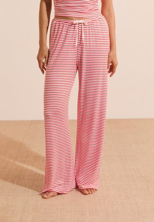 Femme portant un pantalon large à rayures horizontales rouges et blanches avec une taille à cordon, debout pieds nus sur un sol recouvert de moquette beige.