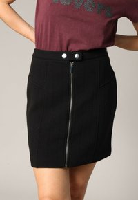 Jupe mini noire en tissu texturé, fermeture éclair avant et deux boutons-pression argentés à la taille. Elle comporte des poches latérales et des détails de couture verticales.