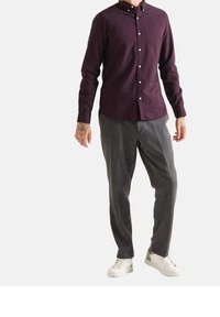 Uomo che indossa una camicia button-up di un viola scuro, pantaloni grigi, sneakers bianche, con tatuaggi visibili su entrambe le mani, su uno sfondo bianco.