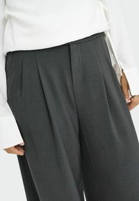 Gros plan d'une personne portant un pantalon gris foncé sur mesure avec des plis et un chemisier blanc à manches longues avec des poignets allongés.