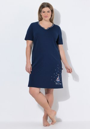 Frau steht barfuß und trägt ein marineblaues, kurzärmeliges Kleid mit einem Segelboot-Design und dem Schriftzug "Take me away" in der Nähe des Saums.