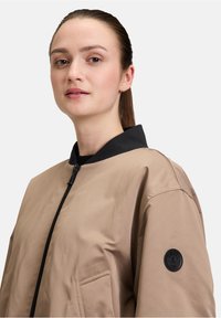 Giacca bomber marrone chiaro con colletto nero e chiusura a zip. Presenta una toppa rotonda nera sul manica sinistra e una tasca laterale.