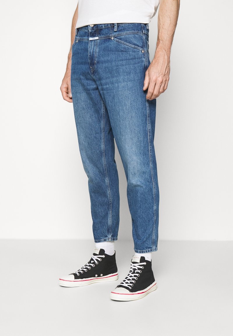 Homme portant un jean bleu court, des chaussettes blanches, des baskets montantes noires et une chemise blanche, debout devant un fond clair uni.