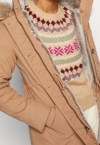 Světlá hnědá parka s podšívkou z umělé kožešiny, předním zipem a stahováním v pase na šňůrku. Pod ní pletený béžový svetr s vícebarevnými geometrickými vzory.