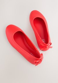 Paar rote Ballerinas mit abgerundeten Zehen und kleinen dekorativen Schleifen auf der Rückseite, von oben auf hellem Hintergrund gezeigt.