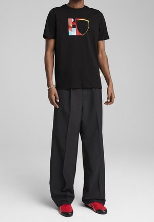 Mand iført sort t-shirt med farverigt geometrisk skjoldlogo, brede sorte bukser, røde og sorte sko og sølvarmbånd på venstre håndled.