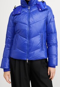 Modrá prošívaná puffer bunda s předním zipem, vysokým límcem, patkami na ramenech se zapínáním na snap knoflíky a bočními kapsami na zip, nosí se s černými kalhotami.