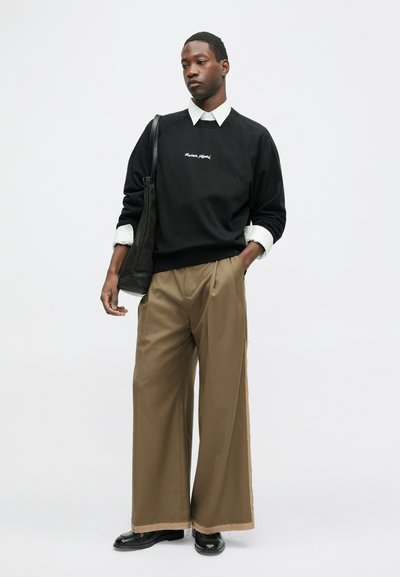 Mustaga sviiter, millel on kirjalik logo, valge särk all, laiad khaki püksid ja mustad kingad. Lihtne kott üle õla.