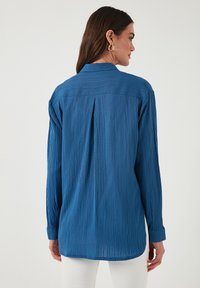 Langärmliges, zerknittertes blaues Hemd aus leichtem Stoff. Mit einem Rückenfalten und geknöpften Manschetten, sowie einem abgerundeten Saum.