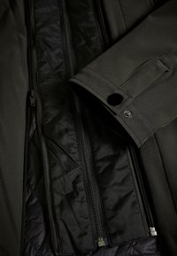 Manche d'une veste noire avec un poignet à bouton-pression reposant sur une doublure intérieure matelassée noire partiellement dézippée avec des fermetures éclair visibles.