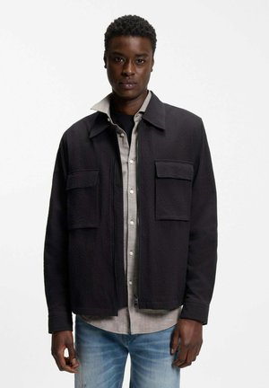 Uomo che indossa giacca nera con cerniera e due tasche sul petto, camicia beige abbottonata sotto e jeans blu sbiaditi, in piedi su sfondo bianco.