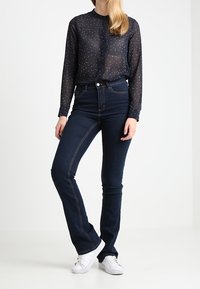 Långärmad, genomskinlig blus i kolagrå med vita prickar, matchad med högmidjade mörkblå utsvängda jeans och vita sneakers.