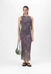 Μωβ εφαρμοστό φόρεμα με σχέδιο κυμάτων, διάφανο ύφασμα και στρογγυλή λαιμόκοψη. Το μοντέλο κρατά μια μεταλλική ασημένια τσάντα και φορά μαύρες πλατφόρμες.
