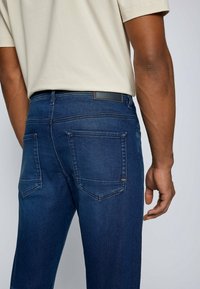 Man med mörkblå denimjeans och en beige kortärmad skjorta, sedd från midjan och nerifrån bakifrån.