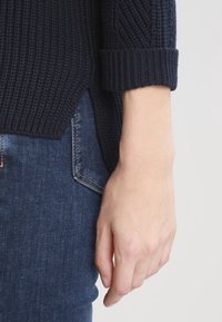 Navy knitted sweater with a textured pattern and rolled cuffs, paired with blue denim jeans. Close-up of hand resting on jeans.