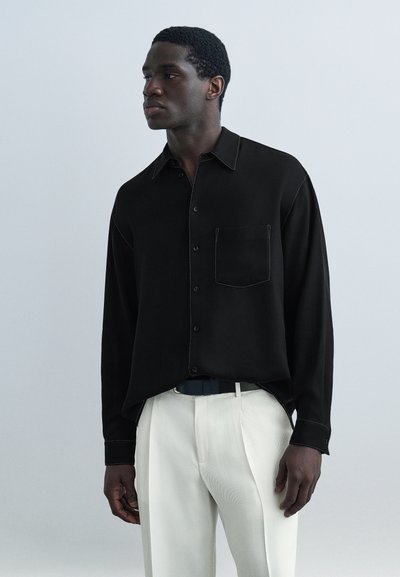Homme portant une chemise noire à manches longues avec boutons et poche avant, rentrée dans un pantalon blanc taille haute, debout devant un fond uni.