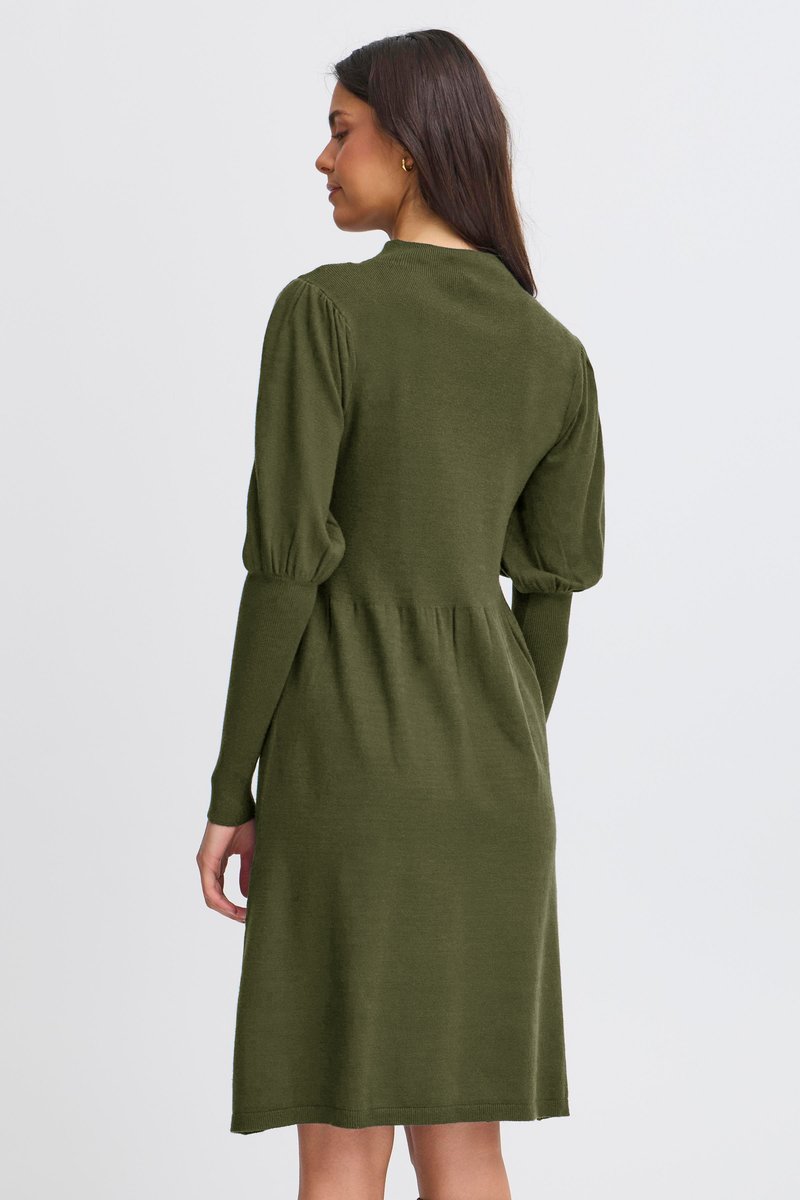 Femme aux longs cheveux foncés portant une robe en maille vert olive avec des manches bouffantes, vue de dos sur un fond uni.