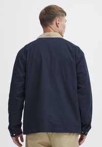 Marinblå bomullskappa med en beige krage. Har långa ärmar och knappade manschetter, strukturerad tyg och en avslappnad passform.