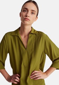 Blusa verde leggera con colletto, maniche a metà e scollatura a V. Presenta un discreto dettaglio di cucitura lungo la parte centrale. Tessuto liscio.