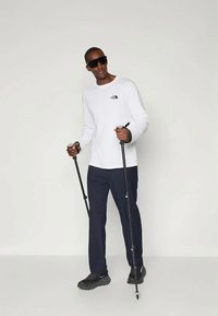 Biała koszula z długim rękawem z czarnym logo, granatowe spodnie i czarne buty. Trzyma kijki trekkingowe, nosi ciemne okulary przeciwsłoneczne. Prosty design, odzież sportowa.