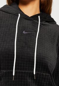 Černá mikina s kapucí vyrobená z texturované tkaniny, s bílým šňůrkovým stahováním a malým černým vyšívaným logem Nike na hrudi.