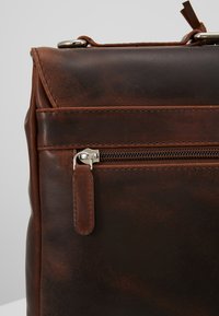 Brun lædertaske med en glat overflade, der har en frontlomme med lynlås og metalhardware. Struktureret design med synlige syninger.