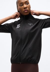 Γυναίκα που ρυθμίζει το γιακά ενός μαύρου ζακέτας με φερμουάρ, με τα λογότυπα "WORLD SERIES OF FITNESS RACING" και "HYROX Puma" στο μπροστινό μέρος.