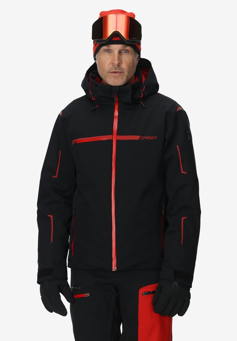 Schwarze Skijacke mit roten Akzenten, mit Reißverschlusstaschen, verstellbarer Kapuze und elastischen Bündchen; kombiniert mit schwarzen Handschuhen und einer Skibrille.
