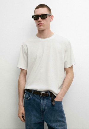 Jonge man met zwarte rechthoekige zonnebril, simpel wit T-shirt met korte mouwen, gevlochten bruine leren riem en blauwe jeans, staande tegen een witte muur.