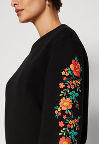 Černá mikina s květinovou výšivkou v živých odstínech oranžové, žluté a tyrkysové podél levého rukávu; měkká textura a kulatý výstřih.