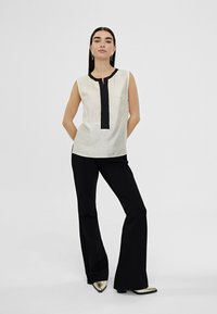 Top sin mangas color crema con tapeta frontal negra, combinado con pantalones acampanados negros y zapatos metálicos de punta. Diseño sencillo y tejido texturizado.