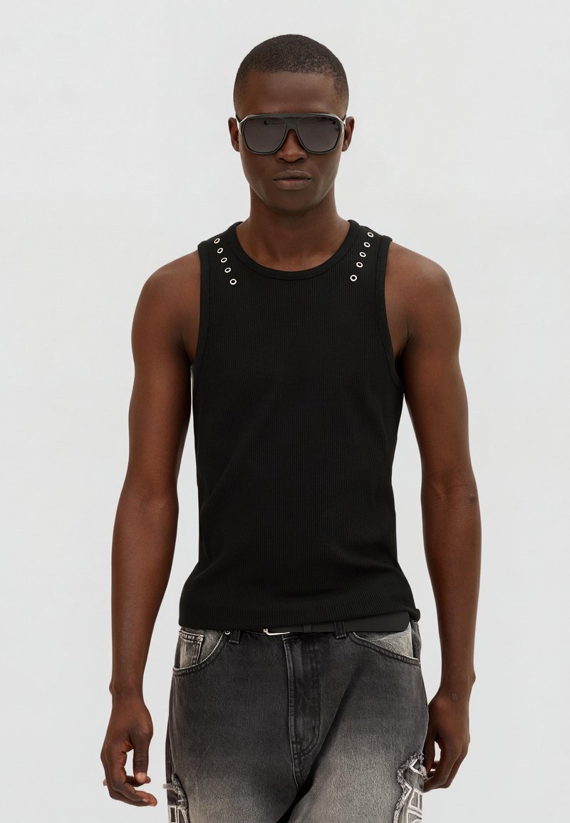 Mężczyzna w czarnym, bezrękawnym topie z prążkowanej tkaniny z oczkowymi detalami przy ramionach, oversize'owych czarnych okularach przeciwsłonecznych oraz wyblakłych czarnych dżinsach z przetarciami.