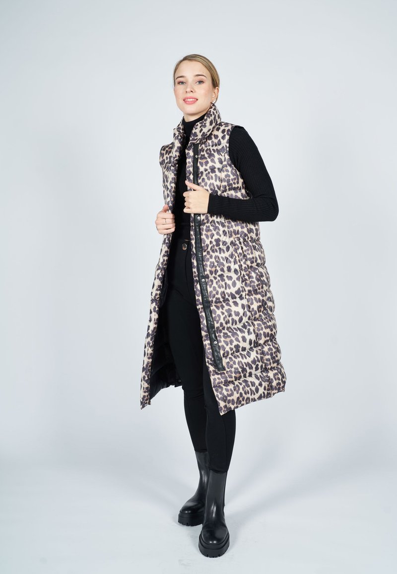 Langer, gepolsterter Weste in Beige mit schwarzem Leopardenmuster. Mit hohem Kragen, Frontreißverschluss und Seitenreißverschlusstaschen. Getragen über einem schwarzen Oberteil und einer Hose.