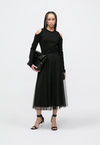 Melns auksts plecu topiņš kombinācijā ar melnu tiula svārkiem ar punktiņiem. Modelis tur melnu somiņu rokā un valkā melnas potīšu siksniņu kurpes.