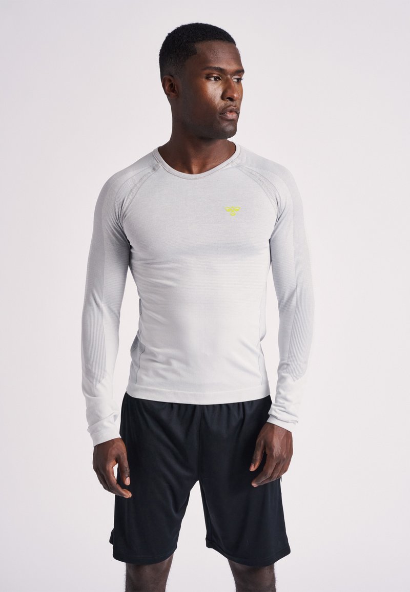 Maglia sportiva a maniche lunghe grigio chiaro con texture a coste e scollo rotondo. Dettaglio logo giallo sul petto, abbinata a pantaloncini neri.