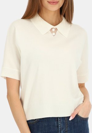 Kobieta ubrana w biały, krótki sweter z dzianiny z ostrym kołnierzykiem i dwoma okrągłymi guzikami przy dekolcie, zestawiony z niebieskimi dżinsami.