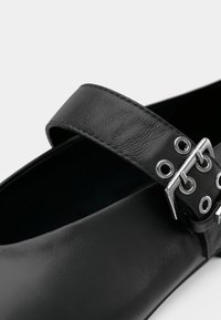 Μαύρο δερμάτινο παπούτσι με φαρδιά λουράκι που διαθέτει ασημένια εξαρτήματα και λεπτομέρειες με κρικάκια. Ομαλή υφή και στρογγυλεμένο σχήμα με κρυφό μύτο.