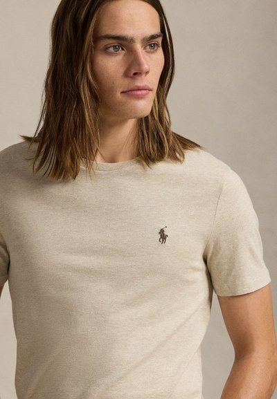 Mladý človek s hnedými vlasmi po ramena, ktorý má na sebe béžové tričko s krátkym rukávom s malým vyšívaným logom hráča póla na hrudi.