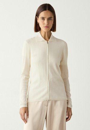 Femme aux cheveux bruns raides porte un cardigan zippé côtelé crème et un pantalon beige, debout devant un fond clair uni.