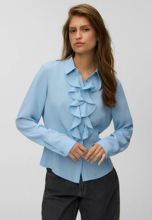 Femme aux longs cheveux bruns portant un chemisier bleu clair avec des volants sur le devant et un pantalon foncé sur un fond uni.