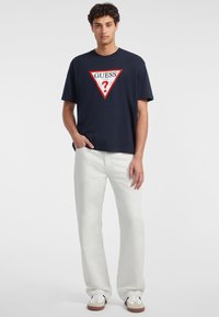 T-shirt in cotone navy con scollo arrotondato, presenta un grafiche triangolare rossa e bianca con il logo "GUESS USA" sul davanti; abbinato a pantaloni color avorio.