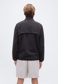 Άτομο με κο ντά ξανθά μαλλιά στέκεται με την πλάτη προς το μέρος μας, φοράει μαύρο μπουφάν με άνοιγμα στην πλάτη και ανοιχτό γκρι σορτς, πάνω σε απλό φόντο.