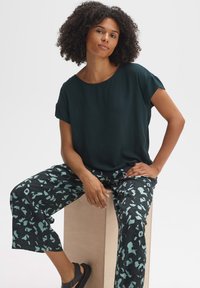 Donkergroene blouse met korte mouwen, gecombineerd met zwarte broek met een mintgroen abstract patroon; model zittend op een licht houten oppervlak.