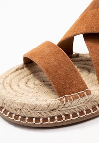 Bruna mocka sandaler med breda remmar och en vävd jutesula, närbild av fram- och sidadetaljer på en vit bakgrund.