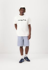Weißes Baumwoll-T-Shirt mit schwarzer Grafik, blaue vertikal gestreifte Shorts, dunkle Sneakers mit hellblauen Streifen und ein cremefarbener Bucket Hat.
