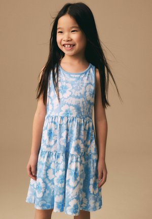 Lächendes Mädchen mit langen schwarzen Haaren, das ein blau-weißes, ärmelloses Kleid mit Blumenmuster vor einem einfarbigen beigen Hintergrund trägt.