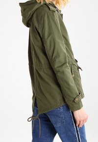 Giacca verde oliva con cappuccio, vita regolabile, dettagli in bottoni e taglio casual. Il tessuto appare liscio con una finitura opaca.