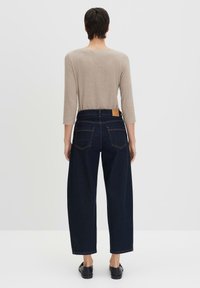 Jeans in denim blu scuro a gamba larga con vita alta, dotati di due tasche posteriori e un'etichetta in pelle marrone sulla vita. Maglietta a maniche lunghe color beige chiaro.