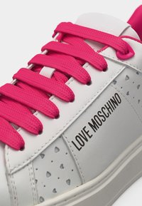 Λευκό αθλητικό παπούτσι με έντονα ροζ κορδόνια, μικρές τρύπες σε σχήμα καρδιάς και το κείμενο «LOVE MOSCHINO» στην πλευρική επιφάνεια.